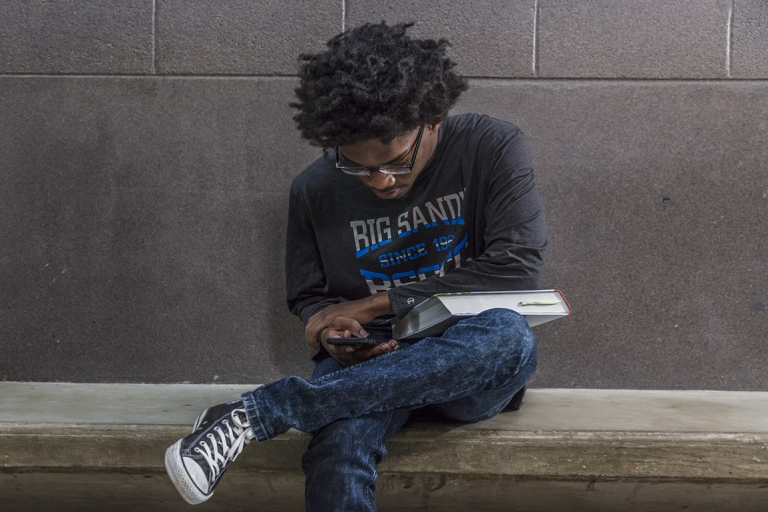 student sitting on a bench looking at their phone and holding a textbook