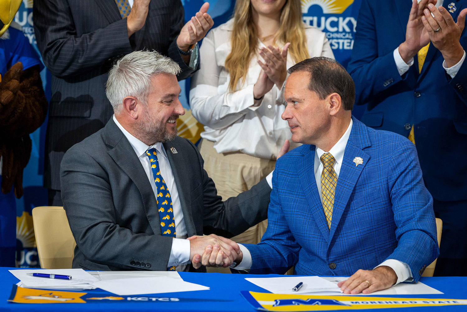 Two men in suits shake hands across a blue table at a formal event, with documents and pens in front of them. The man on the left has white hair and a beard, wearing a dark suit with a white shirt and a blue tie patterned with yellow Kentucky state outlines. The man on the right wears a blue checkered suit, white shirt, and yellow patterned tie. Behind them, a young woman with long blonde hair in a white blouse and beige pants claps, along with others in suits. A partial view of a fox mascot in blue is visible on the left. The backdrop features the Kentucky Community & Technical College System (KCTCS) logo, and table signage includes KCTCS and Morehead State University branding.